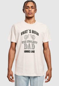 Una camiseta de algodón de color rosa claro presenta un diseño gráfico con el texto "ASÍ ES COMO SE VE EL PAPÁ MÁS GENIAL" y dibujos de pulgares hacia arriba.