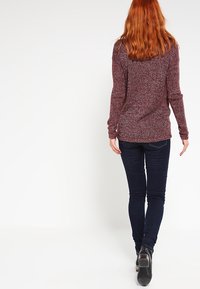 Pull en maille bordeaux avec un design texturé, manches longues et ourlet arrondi, accompagné de jeans skinny bleu foncé et de bottines.