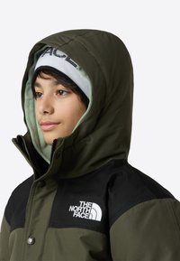 Grønn hettejakke med svarte detaljer og en tydelig hvit logo på brystet, med en mykt foret hette og glidelås foran.