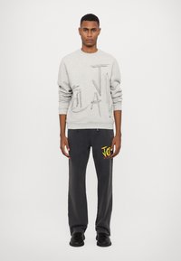 Grå sweatshirt med relieftryck; svarta sweatpants med färgglogotyp; modell stående mot neutral bakgrund; avslappnad urban stil.