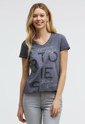 Junge Frau mit langen blonden Haaren, die lächelt, trägt ein blaues grafisches V-Ausschnitt-T-Shirt und hellgraue Jeans, steht vor einem schlichten Hintergrund.