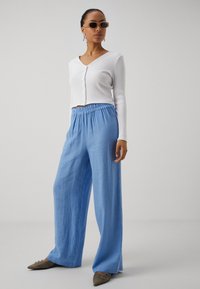 Top court rayé blanc avec des boutons, associé à un pantalon en lin bleu ample et des chaussures marron à talons pointus. Des lunettes de soleil et des boucles d'oreilles complètent la tenue.