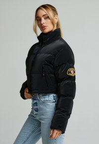 Zwarte cropped pufferjack met een hoge kraag, voorzien van een ritssluiting en een logo patch op de mouw, gecombineerd met lichte denim jeans.