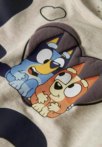 Lysfarget bomull t-skjorte med grafikk av to tegneseriehunder, en blå og en oransje, i en leken posisjon, satt mot en mørk bakgrunn.