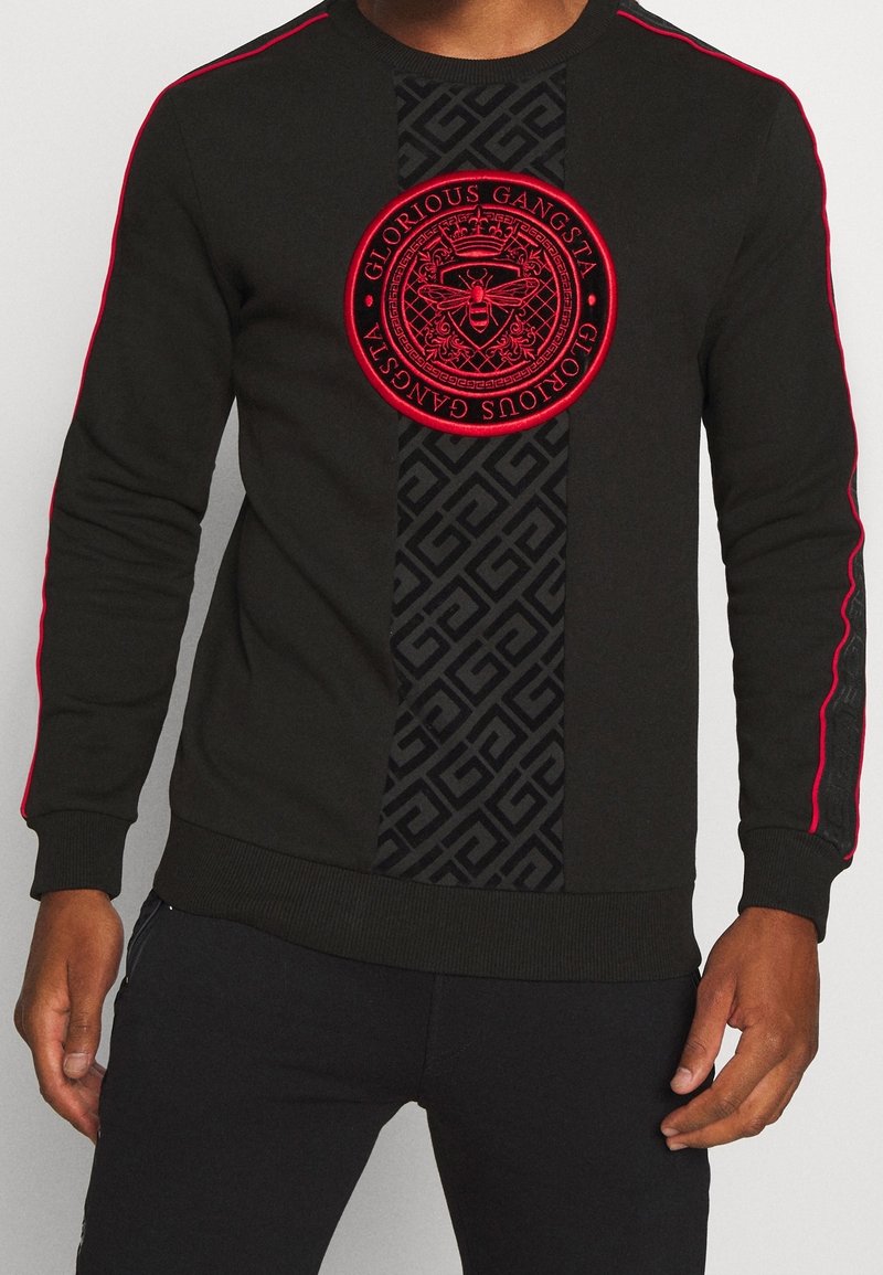 Maglietta nera a maniche lunghe con piping rosso sulle maniche, una striscia verticale a motivo e un emblema circolare rosso con la scritta "Glorious Gangsta" con un design di ape.