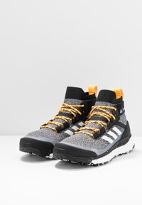 Chaussures de sport noires et grises avec une tige texturée, dotées de lacets orange, de trois bandes blanches proéminentes, et d'une semelle à motif.