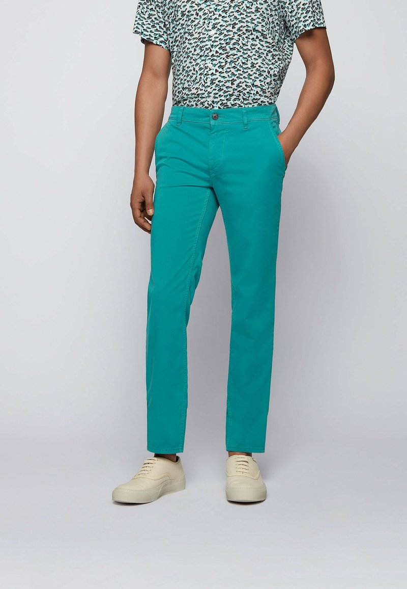 Personne portant un pantalon droit couleur sarcelle, des baskets blanches et une chemise à manches courtes avec un motif abstrait bleu et noir.