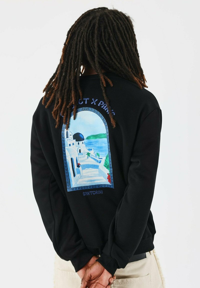 Sudadera negra con un gráfico de una escena de Santorini, con escalones que llevan a edificios blancos y una cúpula azul, y acentos florales rojos.