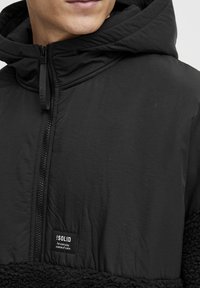 Svart hybridjacka med en slät yttermaterial och mjuk fleece i den nedre delen, med framzipper och logotags.