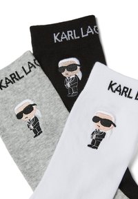 Tři páry ponožek v šedé, černé a bílé barvě, s vyšitým znakem postavy v slunečních brýlích. Text "KARL LAGERFELD" se objevuje na každém páru.