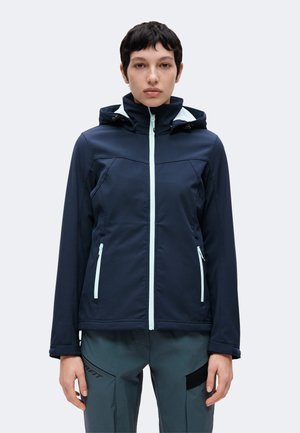 Persona con cabello corto y oscuro, vistiendo una chaqueta con capucha azul marino con cremallera y pantalones gris oscuro, de pie frente a un fondo liso.