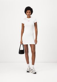 White bodycon dress with short sleeves, ribbed texture, and a pink logo. Paired with a small black bag and chunky silver sneakers.