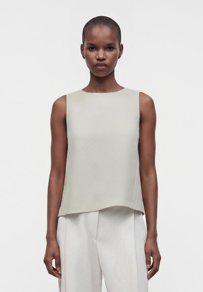 Femme aux cheveux courts portant un haut sans manches beige clair et un pantalon ample assorti, debout devant un fond blanc uni.