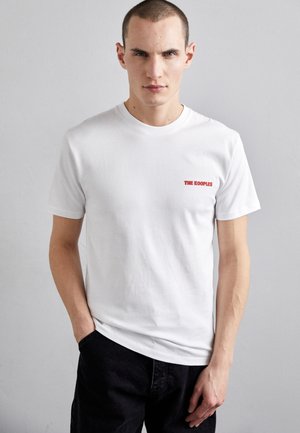 White cotton t-shirt with short sleeves and a crew neckline. Features red text "THE KOOPLES" on the left chest area. Simple design.