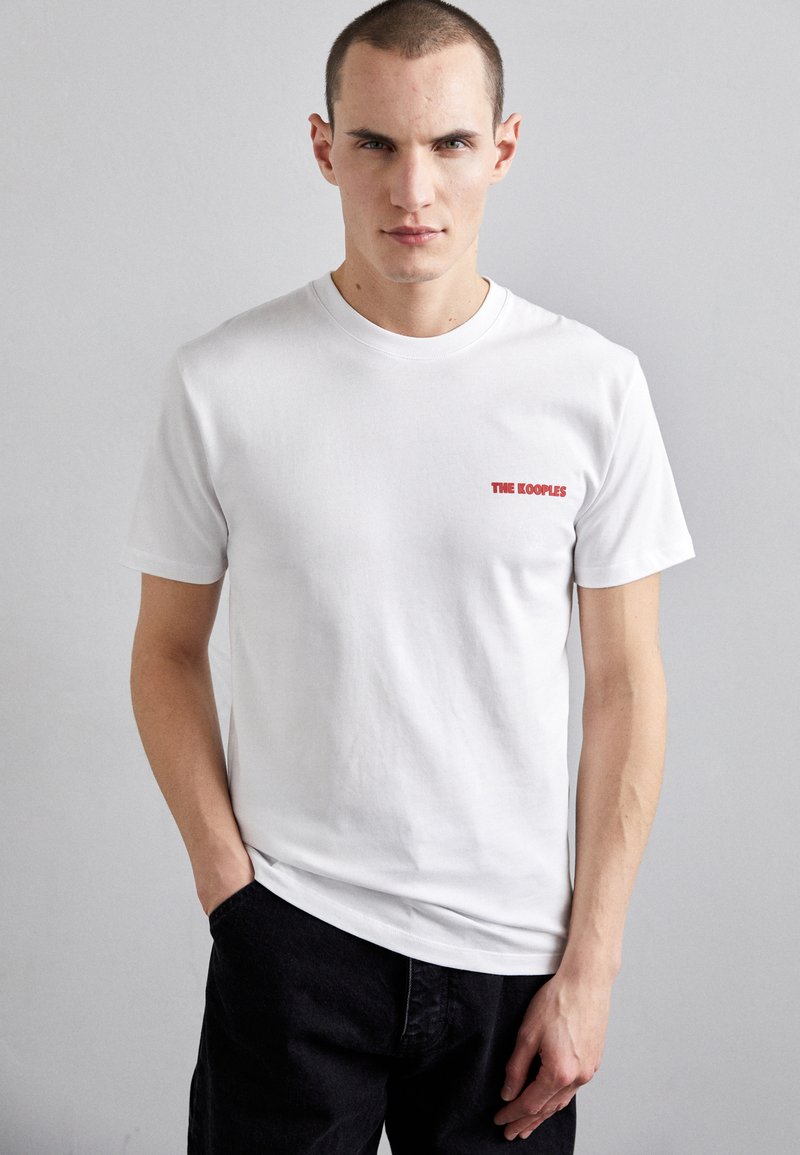 Camiseta de algodón blanca con mangas cortas y cuello redondo. Presenta el texto rojo "THE KOOPLES" en la parte izquierda del pecho. Diseño sencillo.