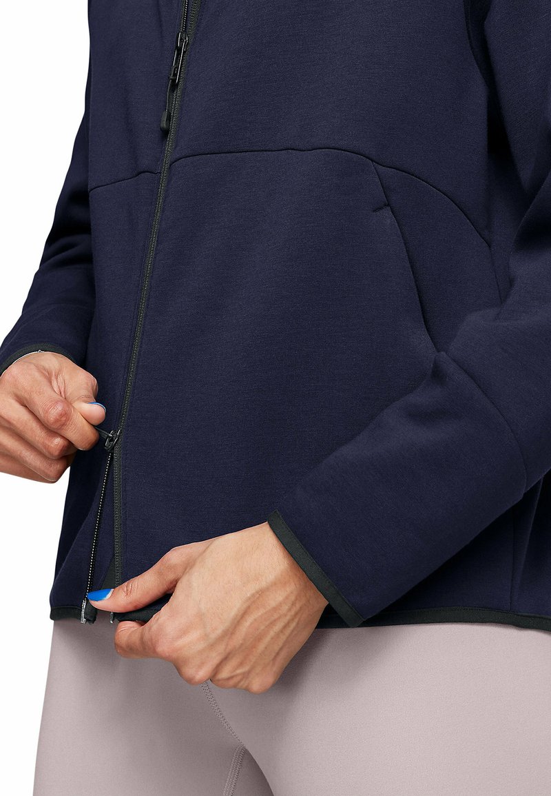 Veste zippée marine avec des finitions sombres, présentant une texture lisse, des poches avant et une main tenant la fermeture éclair, associée à des leggings clairs.
