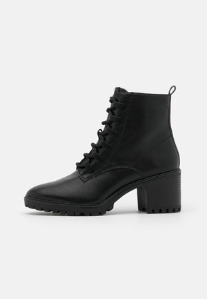 Botte noire en cuir à la cheville avec talon épais, semelle crantée, lacets à l'avant et languette à l'arrière, vue de profil sur fond blanc.