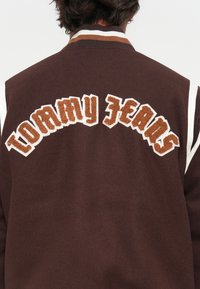 Giacca varsity marrone con accenti bianchi, caratterizzata da testo ricamato in rilievo "TOMMY JEANS" in marrone chiaro e scuro sulla schiena.