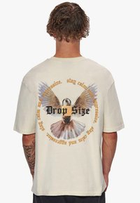 T-shirt en coton beige clair avec un graphisme d'un oiseau aux ailes déployées au dos, entouré de texte en noir et orange.