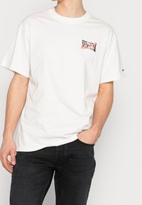Camiseta de algodón blanca con un colorido logo de "TOMMY JEANS" en el pecho, con un corte holgado y mangas cortas. Llevada con jeans oscuros.