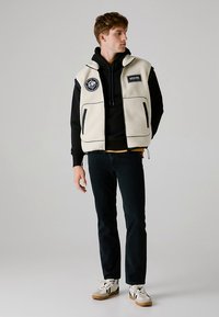 Bež flis brezrokavnik s črnimi poudarki, dvema sprednjima žepoma in logotipom, nošen preko črne kapucarice in temnih kavbojk, s belimi supergami.