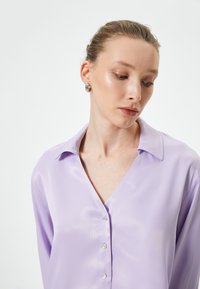 Lavendel satinbluse med V-udskæring, lange ærmer og knaplukning fortil, med en glat og blank overflade samt klassisk krave.
