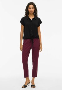 Črna kratkosleeve bluzica s ovratnikom in gumbi, v kombinaciji z bordo slim-fit hlačami. Model nosi črne sandale na peto z odprtim prstom.