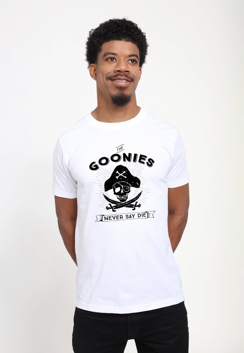 Mężczyzna w białej koszulce z czarną grafiką pirackiej czaszki i mieczy oraz tekstem „The Goonies Never Say Die”, stoi, uśmiecha się, ręce za plecami.