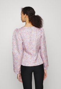 Blusa floral lilás clara com mangas bufantes e punhos elásticos. O padrão apresenta pequenas flores laranjas e amarelas sobre um tecido texturizado.