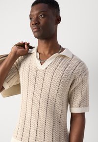 Strick-Poloshirt in Creme mit einem strukturierten Zickzackmuster, kurzen Ärmeln und einem kontrastierenden Kragen. Das Modell hält eine Tasche.