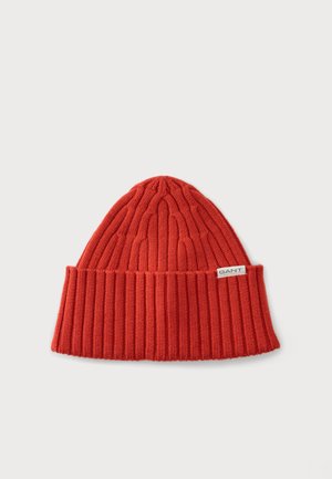 Bonnet rouge en laine avec bord replié et petite étiquette blanche indiquant "GANT" sur un fond clair uni.