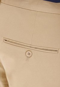 Beżowa tkanina o gładkiej fakturze posiada poziomą tylnią kieszeń z zapięciem na guzik. Szwy są starannie i równo wykonane.