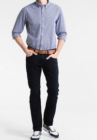 Karierter blau-weißer Hemd, dunkle Jeans, brauer Gürtel und Sneaker mit weißen und olivgrünen Akzenten. Lässige Passform mit hochgeschobenen Ärmeln.