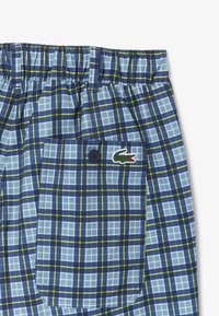Bañador de cuadros azules con cinturilla elástica, un bolsillo tipo parche y el logo de Lacoste. El patrón incluye rayas verdes y de azul más claro.