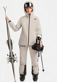 Tenue de ski beige clair, comprenant une veste et un pantalon imperméables. Caractéristiques : capuche, poches zippées et détails noirs. Tient des skis et des bâtons.