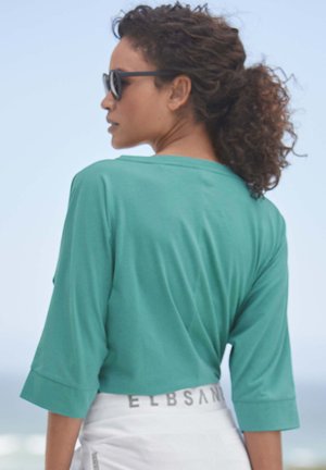 Turquoise, loose-fitting t-shirt with 3/4 sleeves and a rounded neckline. Smooth fabric with a relaxed drape, worn with white shorts featuring a logo.