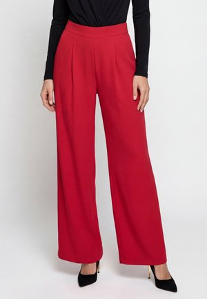 Persona con pantaloni rossi a vita alta e gamba larga abbinati a una maglia nera a maniche lunghe e scarpe nere con tacco alto, in piedi su sfondo bianco.