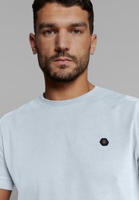 Hellblaues Baumwoll-T-Shirt mit rundem Halsausschnitt und kleinem schwarzen hexagonalen Logo auf der linken Brust. Kurze Ärmel, glatte Textur.