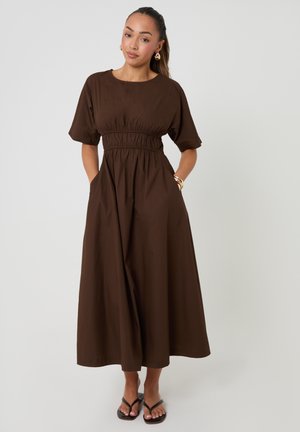 Frau mit zurückgebundenem Haar, die ein braunes Midikleid mit Puffärmeln, tailliert, Hände in den Taschen und schwarze Sandalen vor einem schlichten Hintergrund trägt.