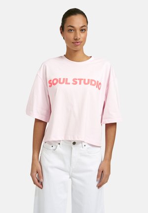 Woman with tied-back hair wearing a light pink oversized T-shirt with "SOUL STUDIO" text and white high-waisted jeans, standing.