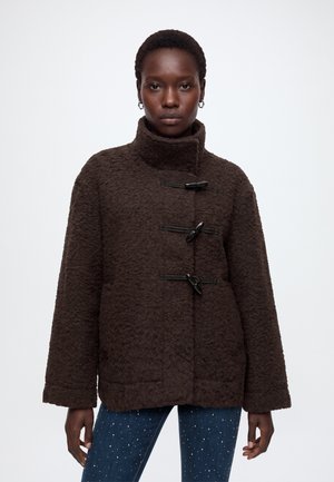 Woman wearing a dark brown textured toggle coat with high collar and blue jeans with small white dots on a white background.