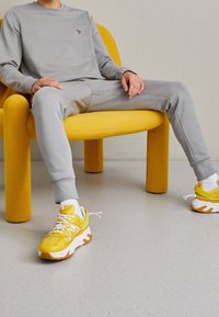Sudadera gris y pantalones de chándal en una figura sentada. Zapatillas deportivas amarillas brillantes con acentos blancos. La silla es de un amarillo sólido, con formas redondeadas.
