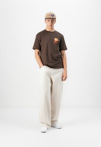 Ung mand iført en brun grafik t-shirt, beige vidde bukser, hvide sneakers og en beige baseball cap, stående mod en hvid baggrund.