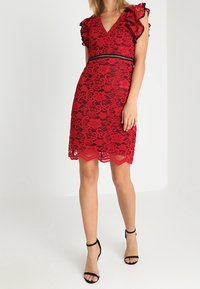 Robe en dentelle rouge avec manches courtes à volants, décolleté en V, ceinture noire, ourlet festonné et motif floral. Portée avec des escarpins noirs à brides.