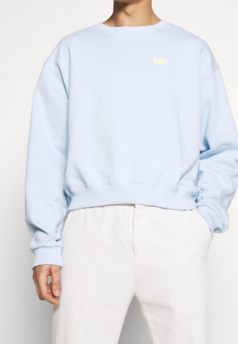 Personne portant un sweat-shirt bleu clair avec un petit logo "MA" et un pantalon en velours côtelé écru, main dans la poche, fond uni.