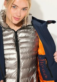 Isolerad jacka med ett metallic silver pufferskikt och ett marinblått ytterlager med orangefärgad foder och grafiska inslag. Dragkedja framtill.