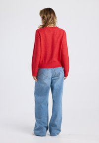 Pull en tricot rouge avec un ourlet côtelé, associé à un jean large bleu clair. Le pull présente un col rond et des manches longues.