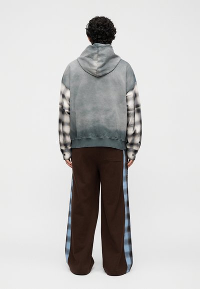 Άτομο που φοράει ξεθωριασμένο μπλε-γκρι φούτερ με κουκούλα πάνω από μαύρο-λευκά καρό μακριά μανίκια και φαρδιά καφέ παντελόνια με μπλε καρό λωρίδες στο πλάι, γυρισμένο από πίσω.