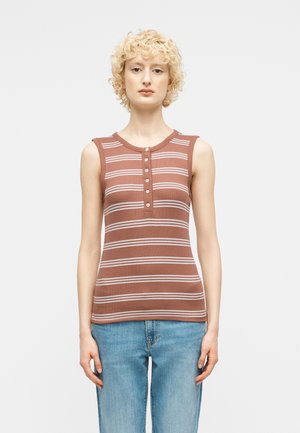 Νέος με κοντά σγουρά ξανθά μαλλιά, φοράει καφέ αμάνικη μπλούζα με λευκές ρίγες και ανοιχτόχρωμο μπλε τζιν, στέκεται προς τα εμπρός.