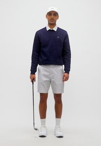 Άνδρας που φοράει μπλε σκούρο πουκάμισο με μακριά μανίκια, άσπρο καπέλο γκολφ, ανοιχτό γκρι σορτς, κρατώντας μπαστούνι γκολφ, στέκεται μπροστά σε απλό φόντο.